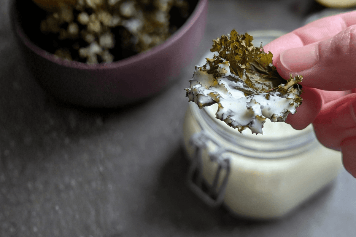 Hand, die einen Grünkohlchip in eine cremige weiße Sauce taucht, über einem Glas, mit einer Schale Grünkohlchips im Hintergrund auf einer dunklen Oberfläche.