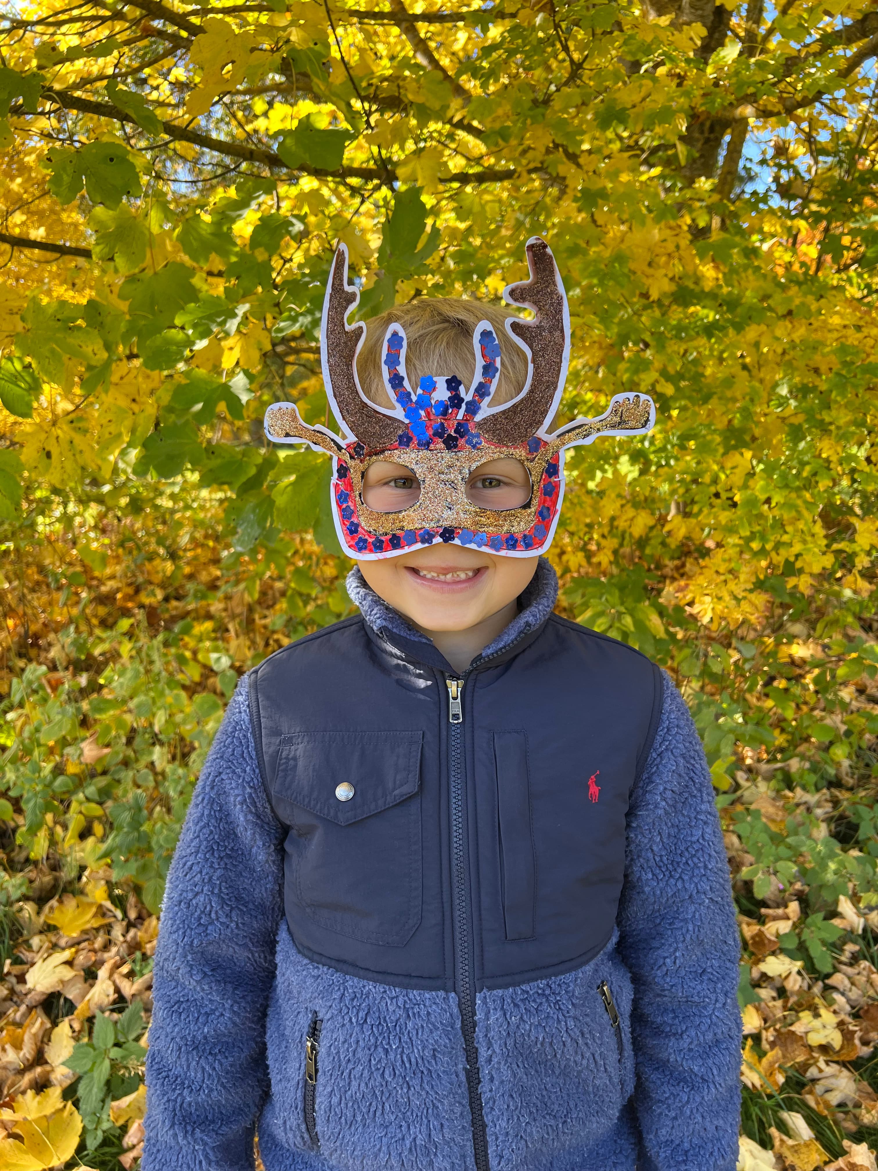 Ein Kind mit einer selbstgestalteten Käfermaske und einer blauen Jacke steht lächelnd vor herbstlichen Blättern.