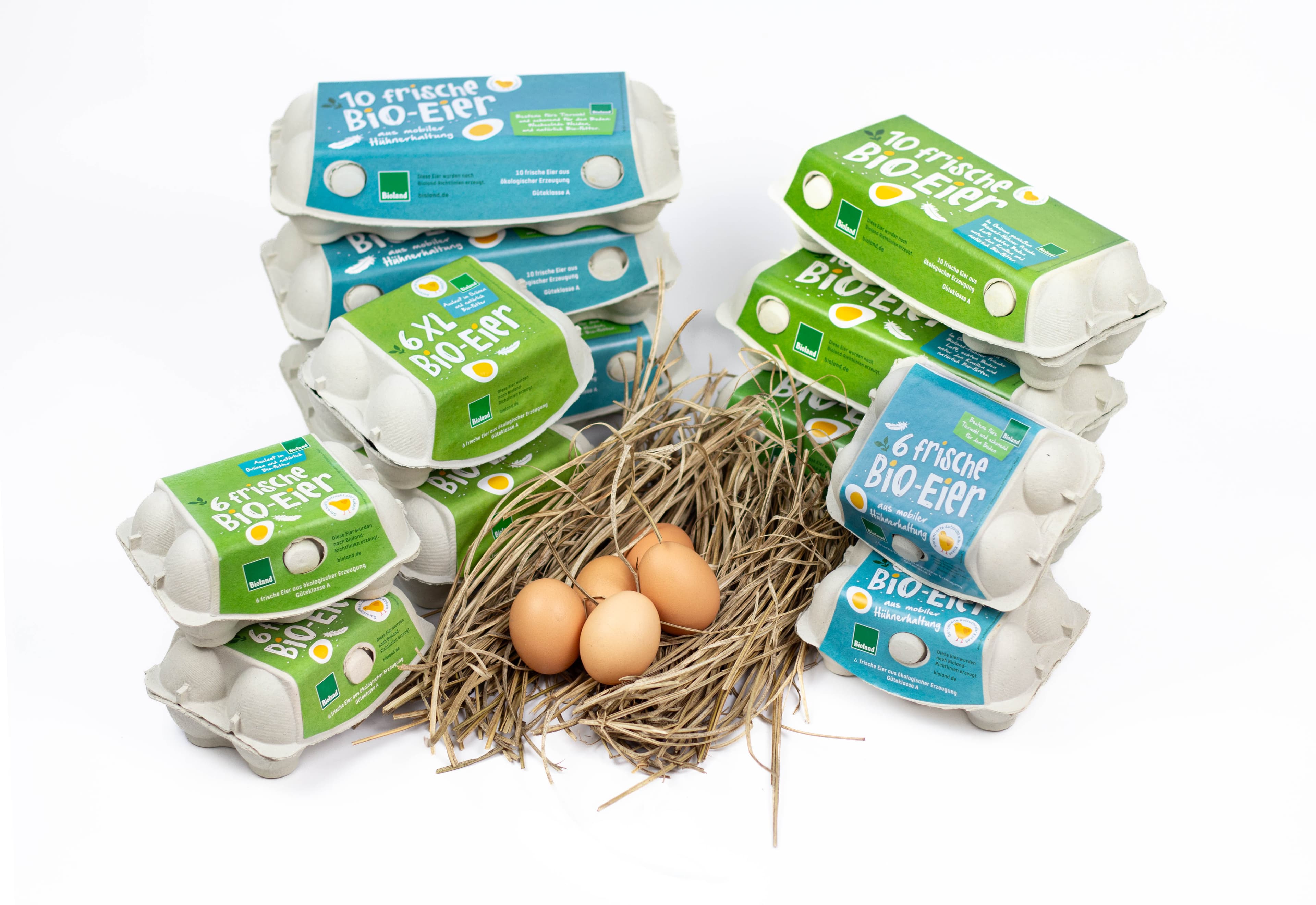 Kartonverpackungen mit Bio-Eiern sind um ein Nest mit drei braunen Eiern auf Stroh gestapelt, vor einem weißen Hintergrund.