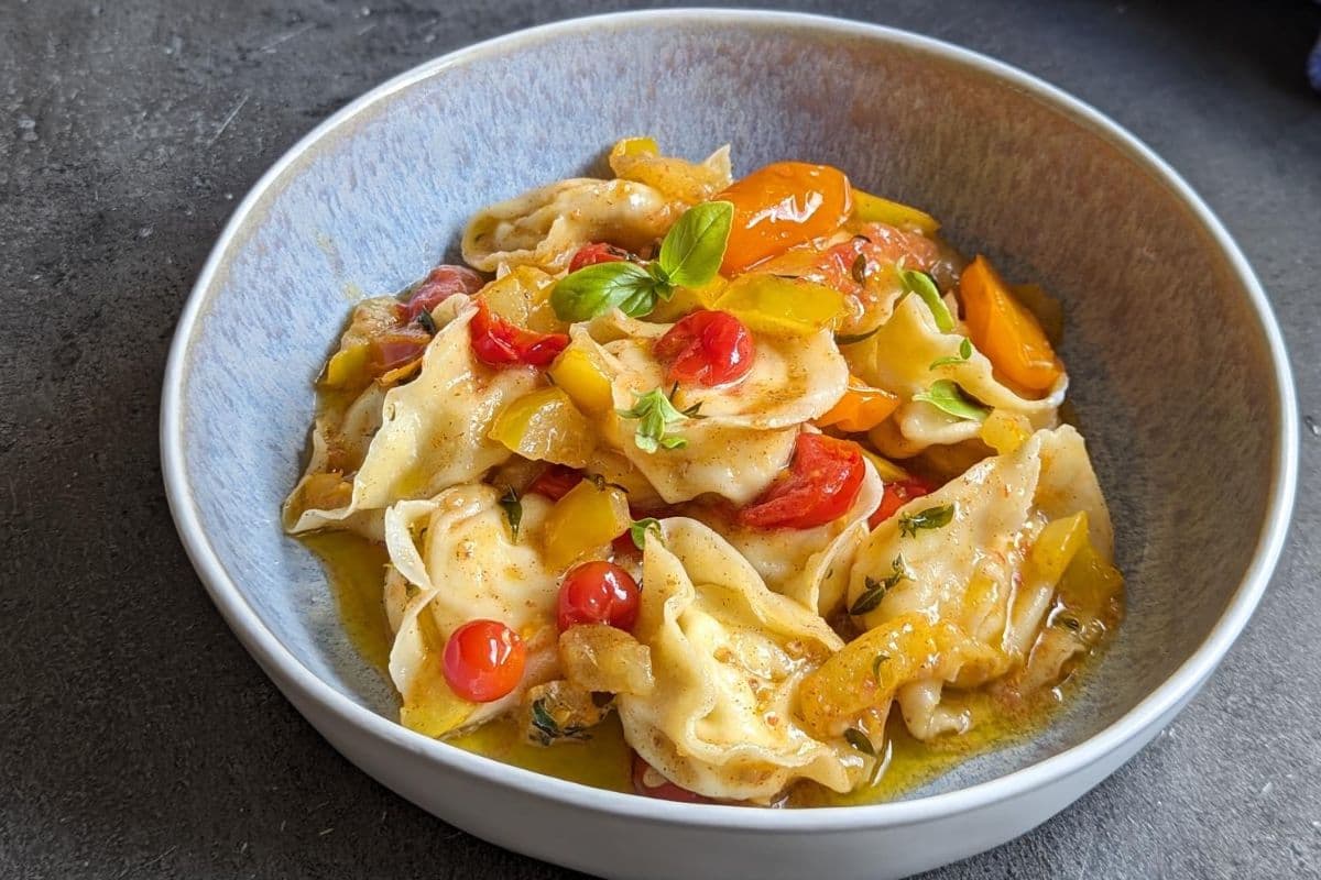 Eine Schüssel Tortelloni mit Kirschtomaten, gelben Paprikaschoten und frischem Basilikum auf einer dunklen Oberfläche.