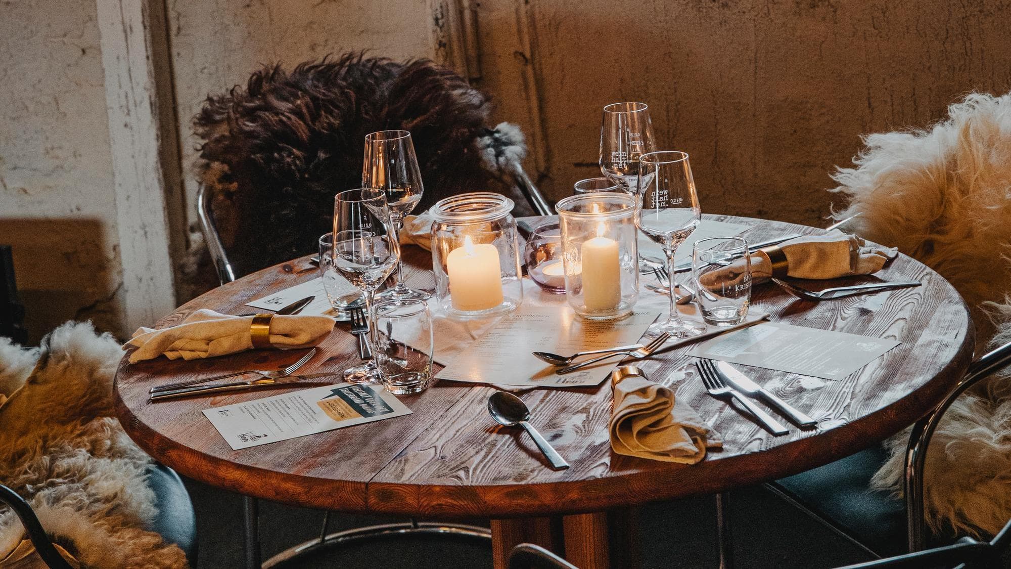Gemütliches Esszimmer-Arrangement mit einem runden Holztisch, Kerzen, Weingläsern, Besteck und mit Fell bezogenen Stühlen, das eine warme, einladende Atmosphäre schafft.