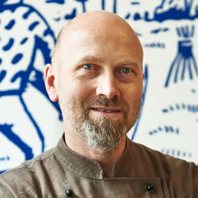 Mann mit Bart, der eine braune Kochjacke trägt und vor einer weiß-blau gemusterten Wand steht.