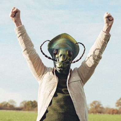 Eine Person mit einer Insektenmaske hebt triumphierend die Arme auf einem Feld unter klarem Himmel.